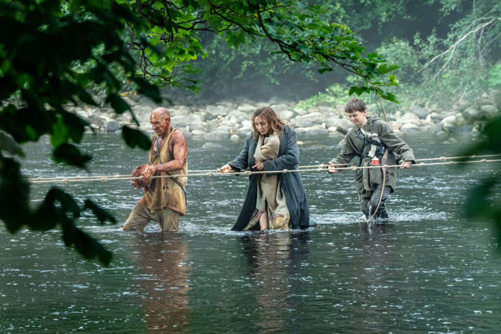 Ralph Fiennes, Jodie Comer, and Alfie Williams crossing a stream